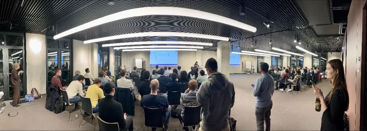 Panoramic view of a packed MLOps Community Munich meetup, attendees seated and standing across a wide room facing the speaker and projected slides.